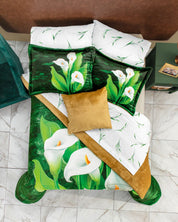 The Juego de Cobertor Supremo Fiori by Intima Hogar features white and green bedding with large calla lily prints, two matching pillows, a plain golden-brown throw pillow, and a floral brown blanket on a white tiled floor.