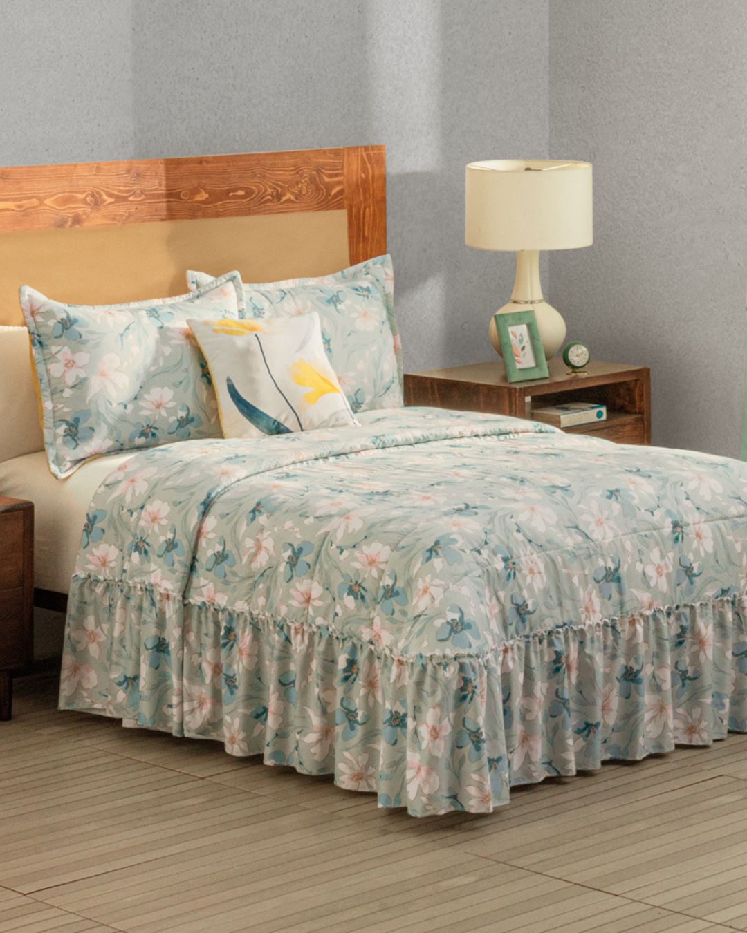 The Juego de Colcha Americana Celina by Intima Hogar, with its light blue floral design and matching pillowcases, paired with a decorative pillow, wooden headboard, and nightstands, creates a cozy and charming bedroom ambiance.