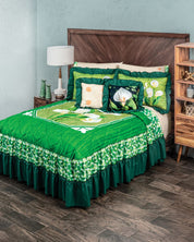 A cozy bedroom features the Juego de Colcha Doble Olán Cala by Intima Hogar—a green floral bedding set with decorative pillows—on a bed with a wooden headboard, next to a white lamp and wooden shelving on dark wood flooring.
