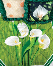 Two decorative pillows—one with button details and one with a calla lily motif—rest on the Juego de Colcha Doble Olán Cala quilt by Intima Hogar, which features white calla lilies and green leaves on a bamboo-inspired background.