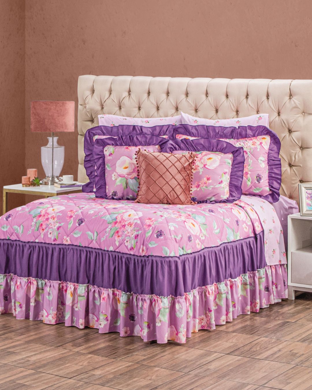 A neatly made bed features the Intima Hogar Juego de Colcha Doble Olán Elin with pink and purple floral patterns, ruffled edges, and decorative shams. A pink lamp and white nightstand sit beside the bed.