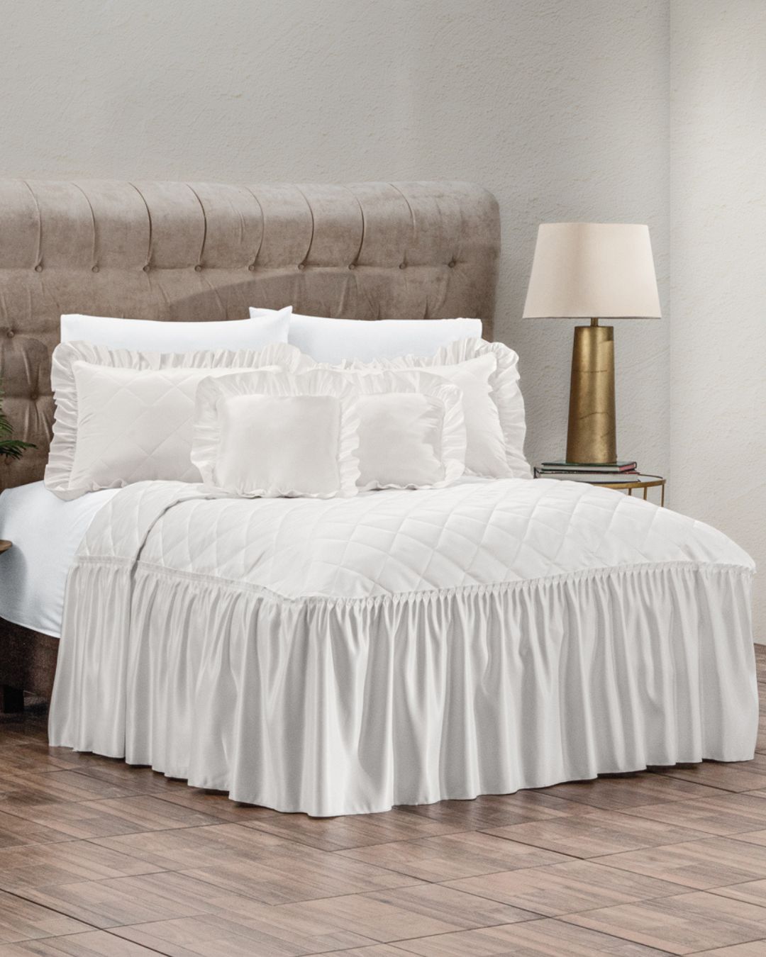 A cozy bedroom features the Juego de Colcha Satinada Paloma by Intima Hogar, set on a beige upholstered headboard with decorative cushions, wood flooring, and a side table topped with a gold lamp and white lampshade.