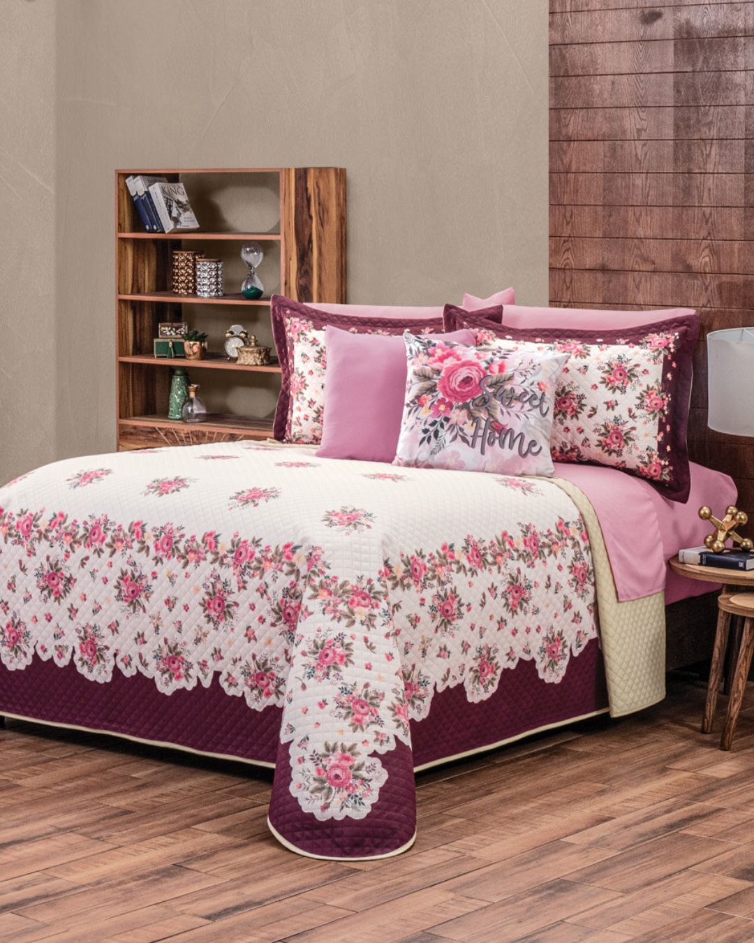 A neatly made bed features the Juego de Colcha Hotelera Yoseline by Intima Hogar in pink, purple, and white with matching pillows. A small bookshelf stands behind, complementing the wooden floors and textured wall.