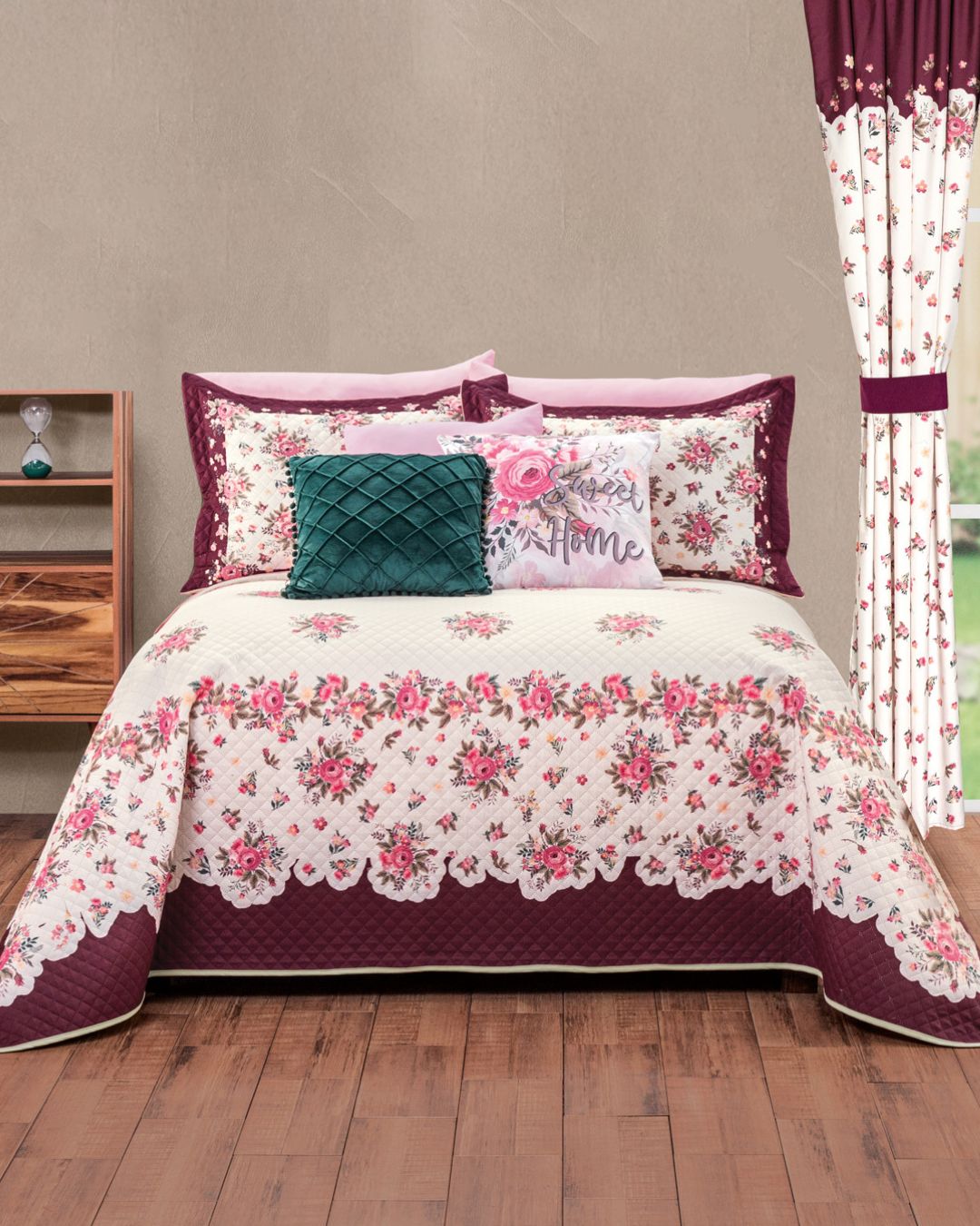 The Juego de Colcha Hotelera Yoseline by Intima Hogar, with floral design and matching pillows in burgundy, pink, and white, is centered in a room with wood floors, a nightstand, and floral curtains by the window.