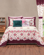 The Juego de Colcha Hotelera Yoseline by Intima Hogar, with floral design and matching pillows in burgundy, pink, and white, is centered in a room with wood floors, a nightstand, and floral curtains by the window.