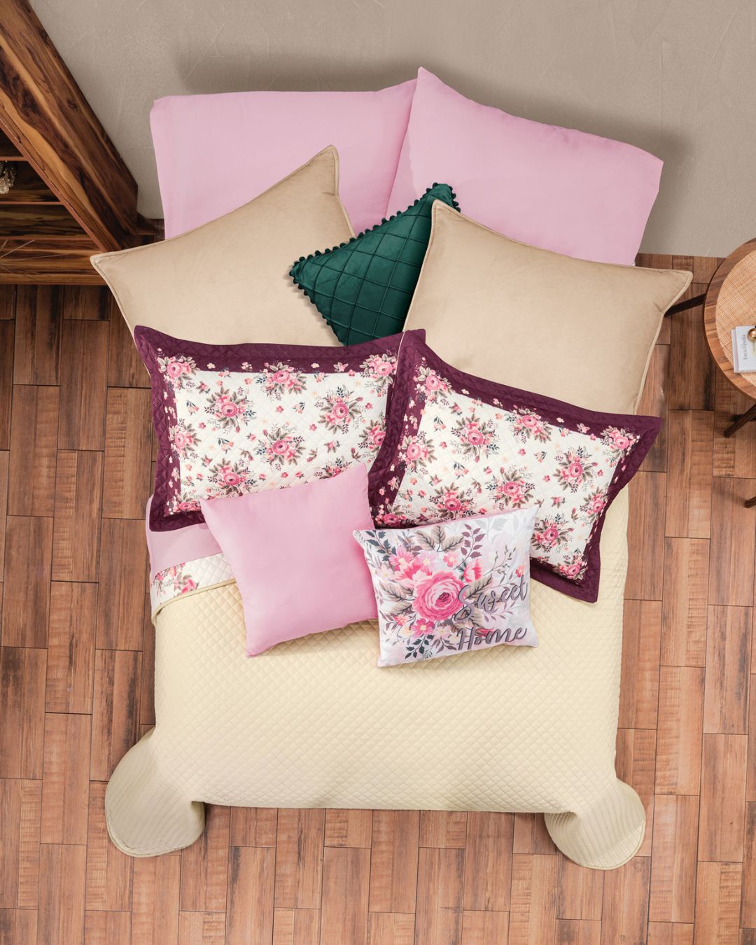 A neatly made bed features the Juego de Colcha Hotelera Yoseline by Intima Hogar, complemented by floral and solid pillows in pink, beige, and green tones, plus a white pillow with pink embroidery, all set on a wooden floor.