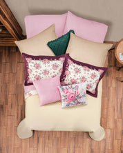 A neatly made bed features the Juego de Colcha Hotelera Yoseline by Intima Hogar, complemented by floral and solid pillows in pink, beige, and green tones, plus a white pillow with pink embroidery, all set on a wooden floor.