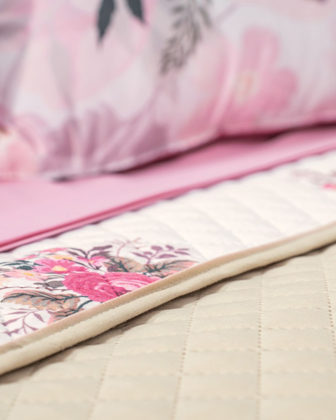 Close-up of a neatly made bed featuring the Juego de Colcha Hotelera Yoseline by Intima Hogar, with beige microfiber, a white and pink floral runner, and matching pink floral bedsheets and pillows in the background.