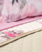 Close-up of a neatly made bed featuring the Juego de Colcha Hotelera Yoseline by Intima Hogar, with beige microfiber, a white and pink floral runner, and matching pink floral bedsheets and pillows in the background.