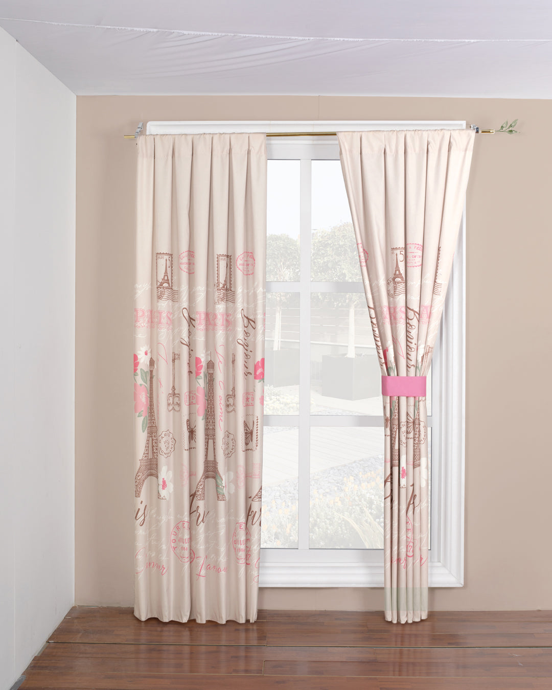 The Juego de Cortinas Ciudad Luz by Intima Hogar, featuring Paris-inspired pink and brown designs, hangs on a window with light brown walls and wooden flooring. One curtain is tied back with a pink band.