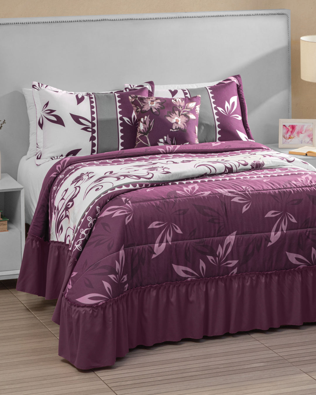 A neatly made bed showcases the Juego de Colcha Americana Parra by Intima Hogar, featuring a purple and white floral comforter set with motivos de follaje, matching pillows including a stylish funda decorativa, and a grey upholstered headboard. Beside the bed on a wooden floor sits a white nightstand with a lamp and small framed picture.