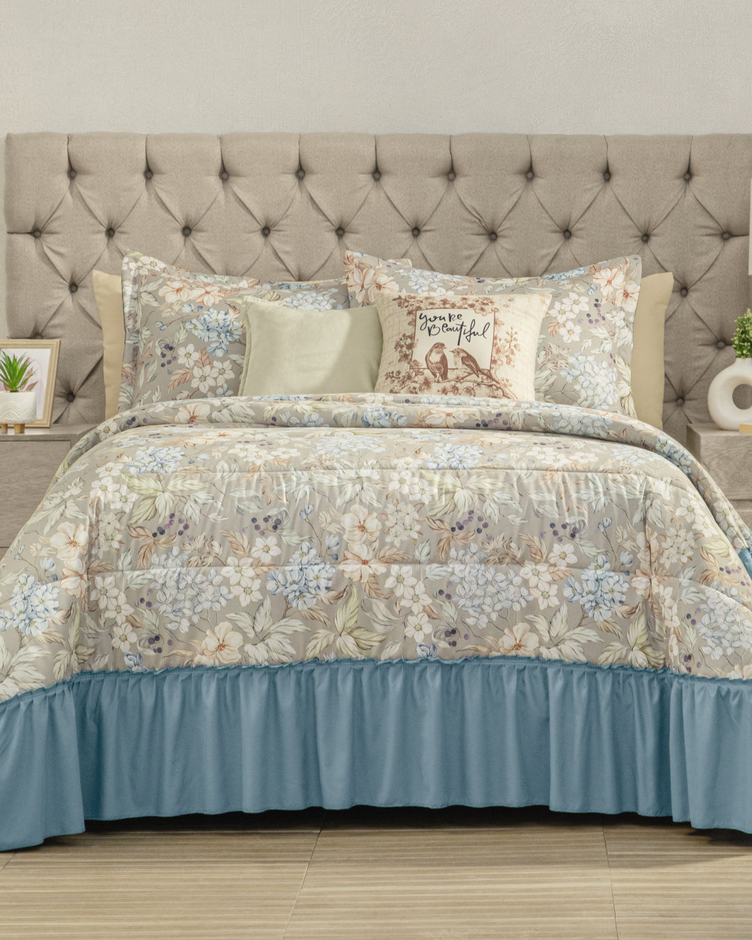 A neatly made bed with the Juego de Colcha Americana Porcelana by Intima Hogar and a light blue bed skirt brings a touch of descanso fresco to the dormitorio. Decorative pillows, including one with text, rest against a tufted beige headboard, while small plants and decor items on the bedside tables complete the inviting look.