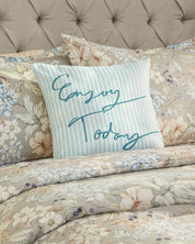 A blue and white striped pillow with “Enjoy Today” written in script sits on a bed dressed in the Intima Hogar Juego de Colcha Americana Porcelana, featuring a floral pattern and an upholstered headboard, creating a dormitorio atmosphere perfect for descanso fresco.