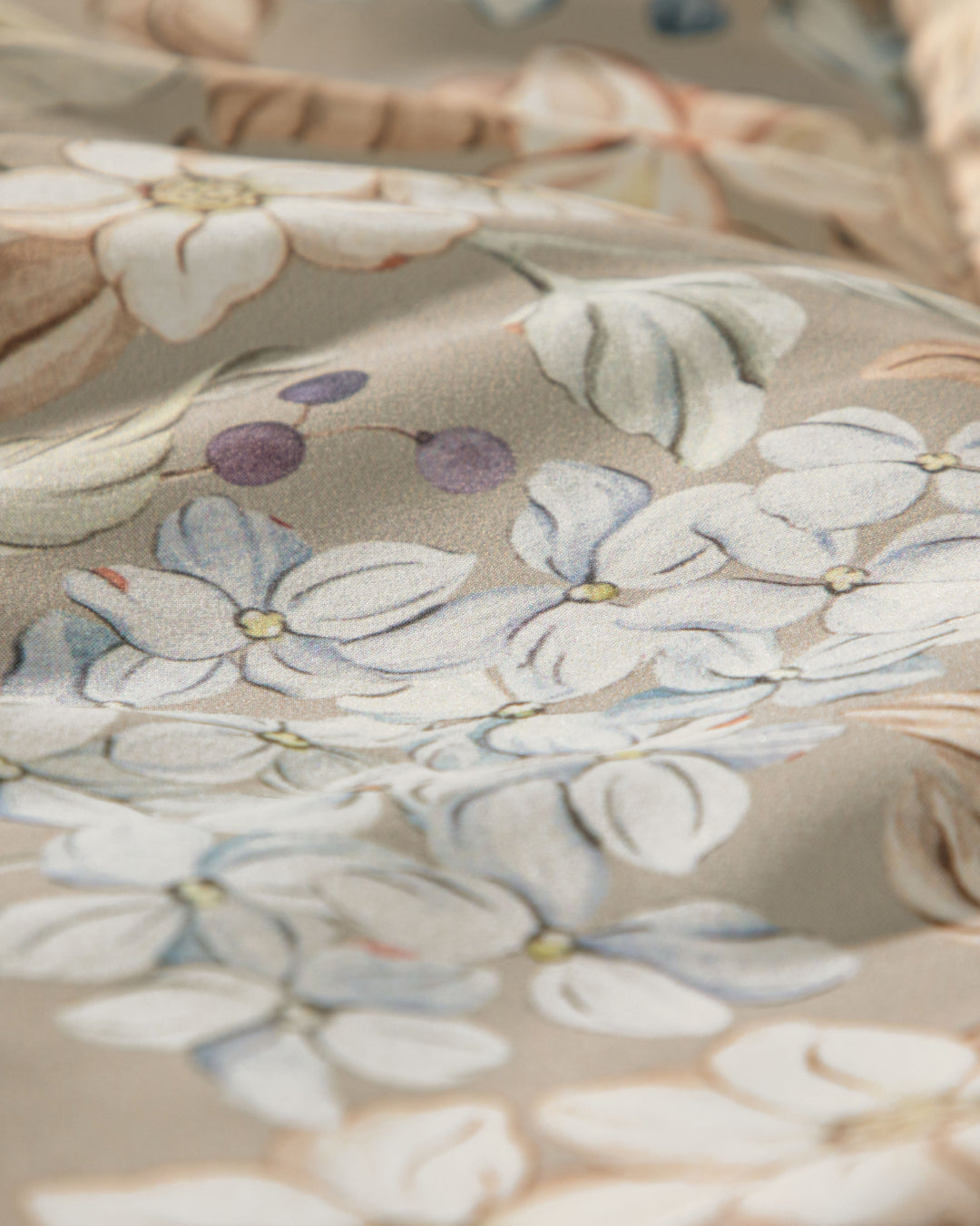A close-up of light-colored fabric with a pastel floral and leaf pattern—featuring blue and white flowers and small purple berries on a beige background—perfect for creating an atmosphere of un descanso fresco in your dormitorio with the Juego de Colcha Americana Porcelana by Intima Hogar.