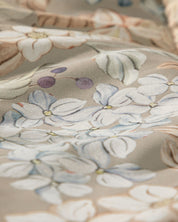 A close-up of light-colored fabric with a pastel floral and leaf pattern—featuring blue and white flowers and small purple berries on a beige background—perfect for creating an atmosphere of un descanso fresco in your dormitorio with the Juego de Colcha Americana Porcelana by Intima Hogar.