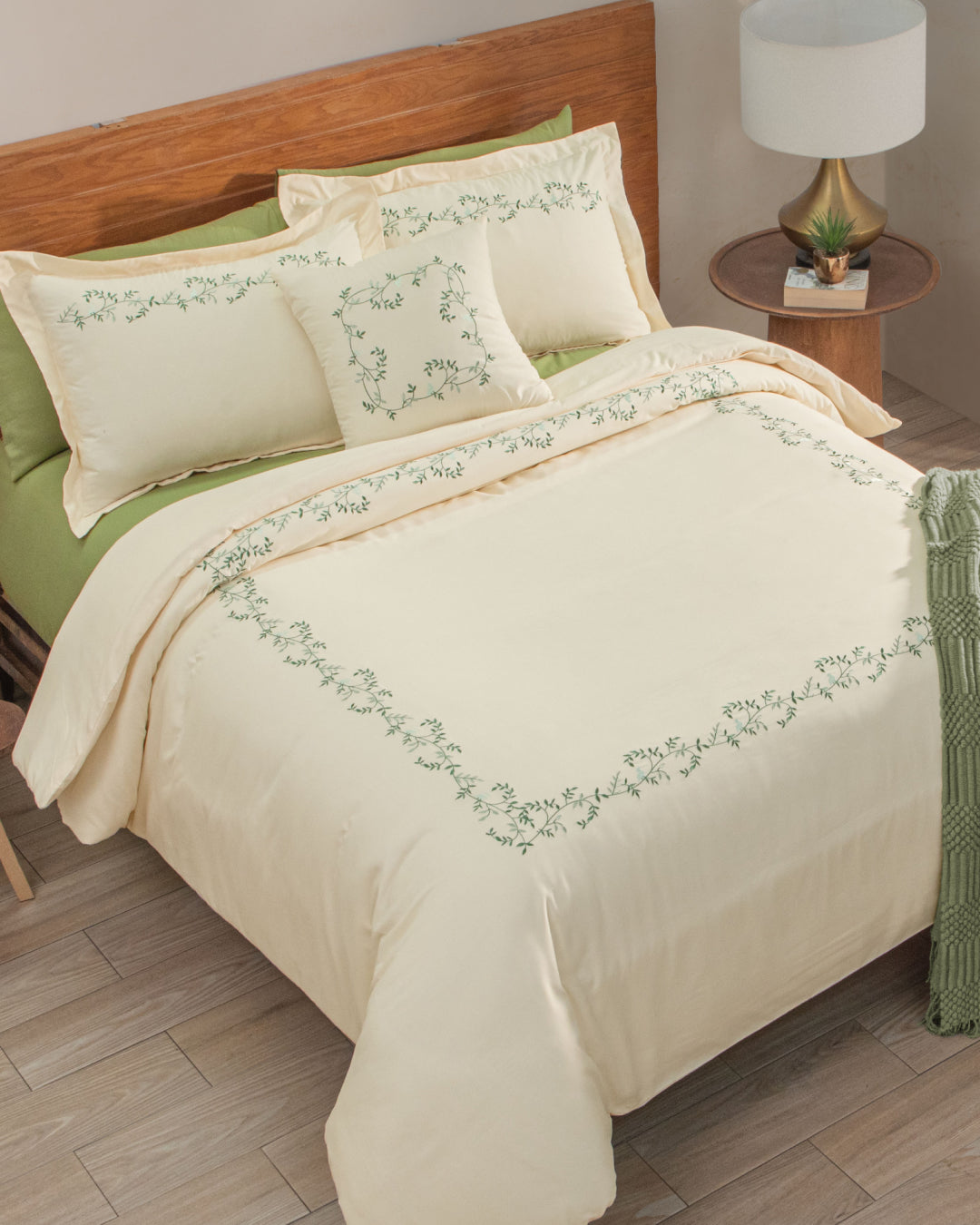 A neatly made bed is dressed in the Juego de Duvet Luxury Marfil by Intima Hogar, showcasing cream-colored bedding with elegant green floral embroidery, complemented by green sheets and matching fundas decorativas pillows. A wooden headboard, bedside table with a lamp, and a small plant complete the scene.