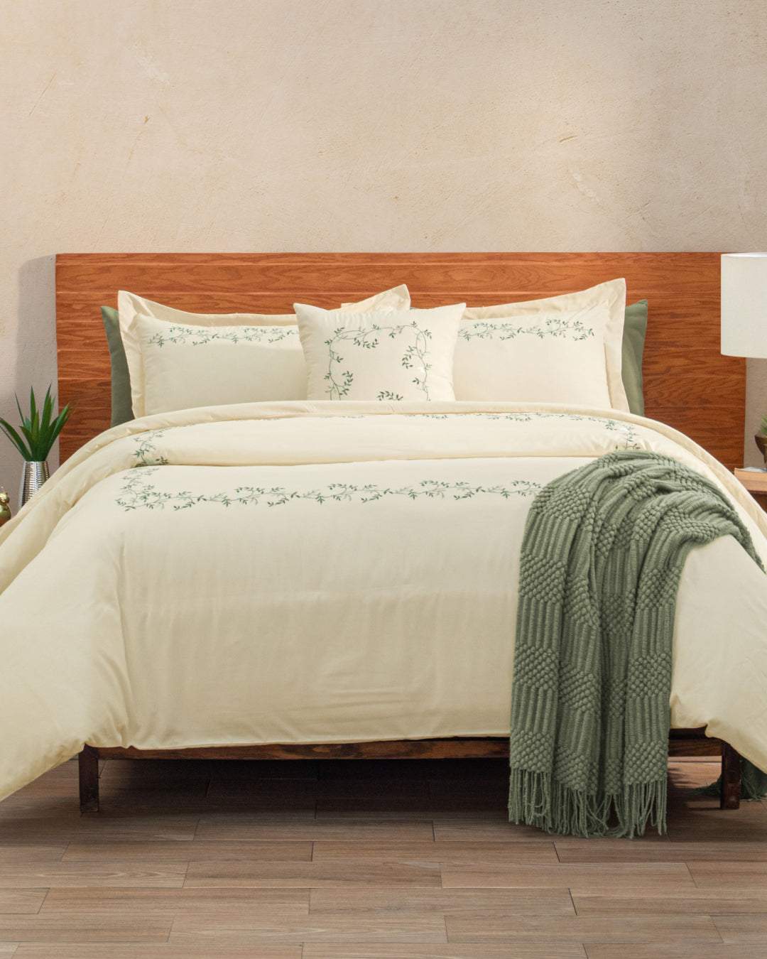 A neatly made bed features the Juego de Duvet Luxury Marfil from Intima Hogar, with cream-colored bedding, green embroidered details, and matching pillows accented by stylish fundas decorativas. A green knit throw is draped over the corner of the edredón blanco. The wooden headboard is complemented by a lamp and plant on the nightstands.