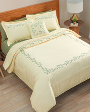 A neatly made bed is dressed in the Juego de Duvet Luxury Marfil by Intima Hogar, featuring light yellow bedding with a green leafy border design and matching fundas decorativas on the pillows, complemented by a green sheet, wooden headboard, round nightstand, lamp, and small potted plant on a wooden floor.