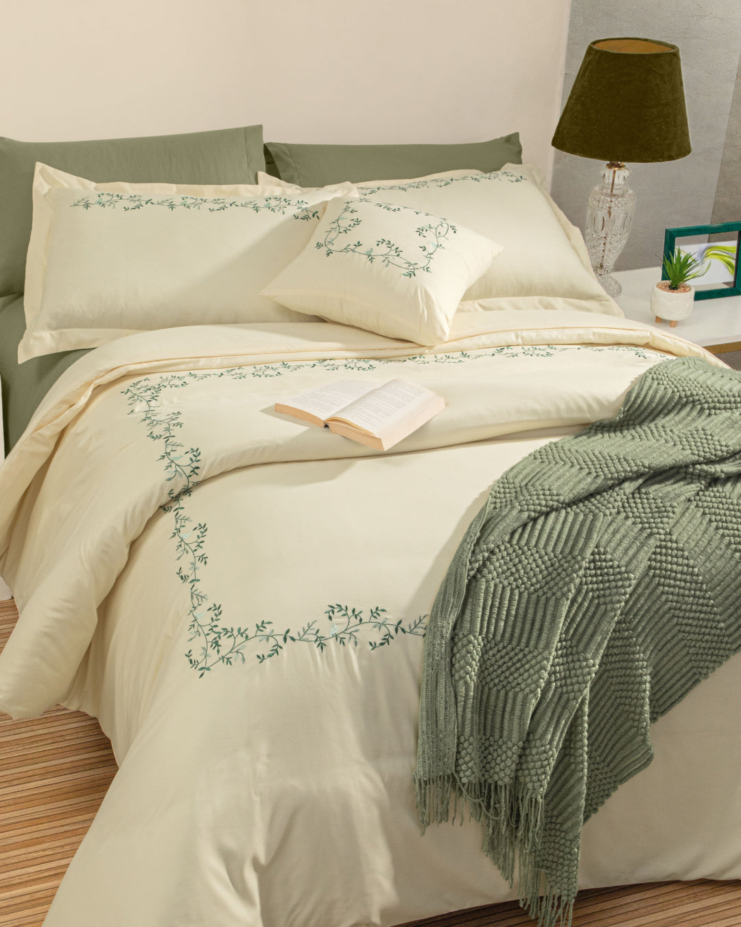 A neatly made bed features the Juego de Duvet Luxury Marfil by Intima Hogar, embroidered with green vines, complemented by green pillows and a textured green blanket. An open book rests beside a lamp on a white nightstand, while fundas decorativas add an elegant touch to this inviting setup.