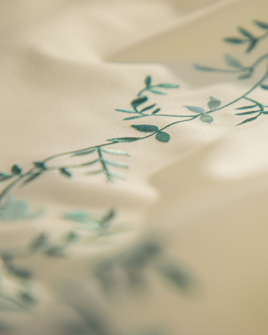 Close-up of cream-colored fabric from the Juego de Duvet Luxury Marfil by Intima Hogar, adorned with a delicate green botanical embroidery pattern of leaves and vines, softly blurred in the background—ideal for complementing an edredón blanco or pairing with elegant fundas decorativas.