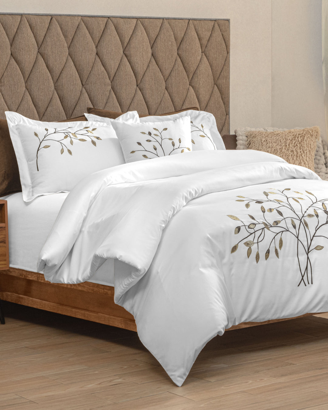 A neatly made bed featuring the Juego de Duvet Luxury Roble by Intima Hogar, showcasing a white duvet with embroidered branches and leaves, two pillows with elegant decorative covers, and a padded, diamond-patterned headboard in a modern bedroom with wood floors.
