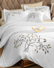 A neatly made bed showcases the Juego de Duvet Luxury Roble by Intima Hogar, a crisp white duvet adorned with embroidered branches and leaves, complemented by two beige decorative pillow covers, and a tray with a cup and saucer resting elegantly on top.