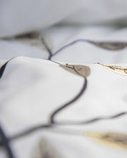 Close-up of the white fabric from the “Juego de Duvet Luxury Roble” by Intima Hogar, with a soft focus, showcasing a pattern of black lines and brown leaves accented with subtle gold details.