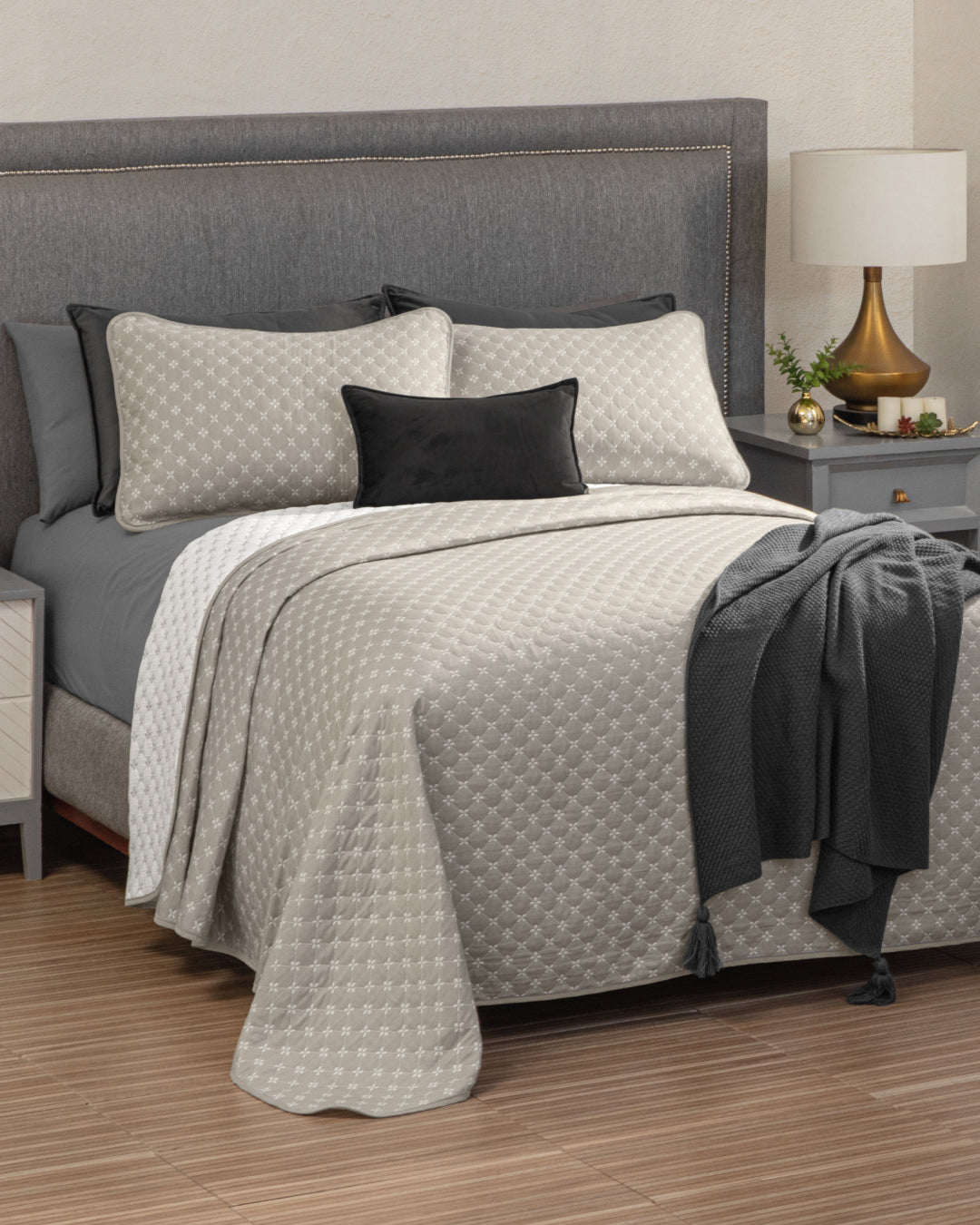 A neatly made bed featuring the Juego de Edrecolcha Doble Vista Laurel by Intima Hogar in beige, paired with a gray upholstered headboard, black and gray accent pillows, a textured gray throw, a bedside table with a lamp, and a small plant.