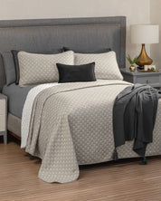 A neatly made bed featuring the Juego de Edrecolcha Doble Vista Laurel by Intima Hogar in beige, paired with a gray upholstered headboard, black and gray accent pillows, a textured gray throw, a bedside table with a lamp, and a small plant.