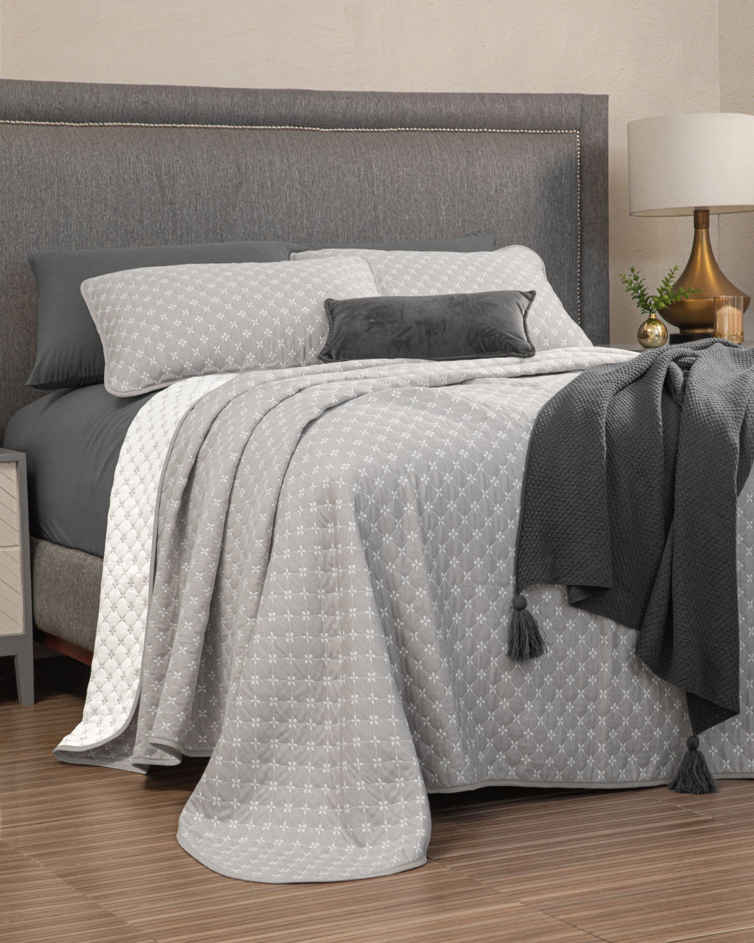 A neatly made bed featuring the Juego de Edrecolcha Doble Vista Laurel from Intima Hogar, complemented by a matching grey headboard, a grey tasseled throw, grey and white pillows, and a bedside table adorned with a lamp and a small green plant.