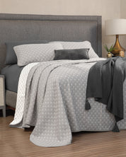A neatly made bed featuring the Juego de Edrecolcha Doble Vista Laurel from Intima Hogar, complemented by a matching grey headboard, a grey tasseled throw, grey and white pillows, and a bedside table adorned with a lamp and a small green plant.