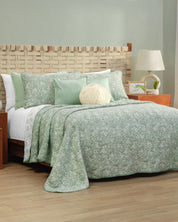Here’s a rewrite of your sentence using the provided product data: The cama is dressed with the Juego de Edrecolcha Doble Vista Olmo by Intima Hogar, displaying sage green and white floral bedding. Four matching fundas decorativas pillows and a round cream cushion complete the look. The woven wood headboard is paired with a nightstand holding a lamp and framed art beside the bed.