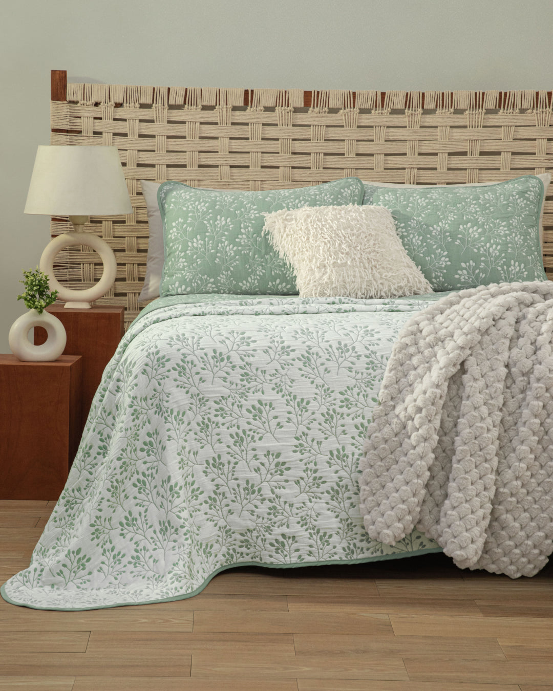 A neatly made cama features the Intima Hogar Juego de Edrecolcha Doble Vista Olmo, showcasing its green and white floral bedding. Two fundas decorativas adorn the pillows, while a fluffy gray colcha is draped over the side. Wooden furniture, a lamp, and a woven headboard complete this cozy and modern bedroom scene.