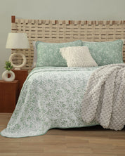 A neatly made cama features the Intima Hogar Juego de Edrecolcha Doble Vista Olmo, showcasing its green and white floral bedding. Two fundas decorativas adorn the pillows, while a fluffy gray colcha is draped over the side. Wooden furniture, a lamp, and a woven headboard complete this cozy and modern bedroom scene.
