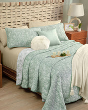 A neatly made cama featuring the Juego de Edrecolcha Doble Vista Olmo by Intima Hogar displays green and white floral bedding, complemented by a round fluffy pillow and a tray with candles on top. Decorative fundas decorativas add charm alongside the wooden headboard, nightstands, and a cream knit blanket to complete this cozy bedroom scene.