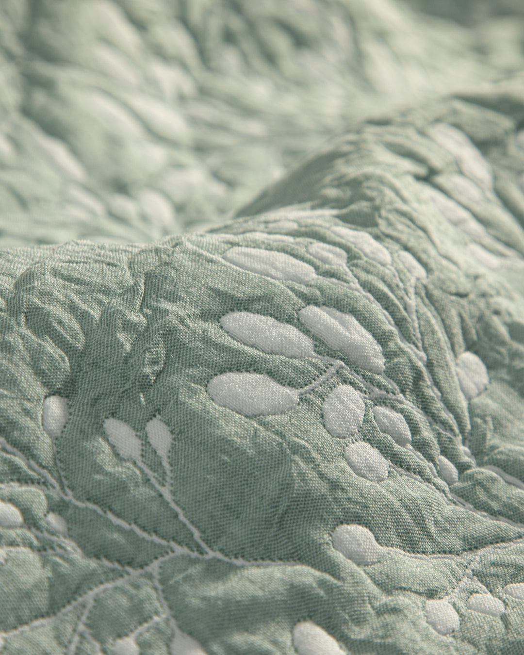 Close-up of the Juego de Edrecolcha Doble Vista Olmo by Intima Hogar, featuring light green quilted fabric with raised white embroidered leaf and branch patterns. The detailed stitching and soft texture make it perfect for a colcha or as fundas decorativas to enhance your cama’s look.