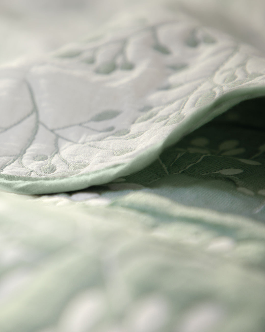 A close-up of the Juego de Edrecolcha Doble Vista Olmo by Intima Hogar showcases its light green fabric, adorned with intricate floral embroidery. The softly folded material highlights the elegant texture and delicate stitching characteristic of this refined bedding set.