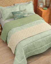 A neatly made bed features the Juego de Edredón Súper Suave Capri from Intima Hogar in green, paired with a pale cream quilted cubrecama and a decorative green pillow with matching fundas decorativas. Beside the bed, a wooden nightstand holds a framed photo, vase, and plant on a light wood floor.