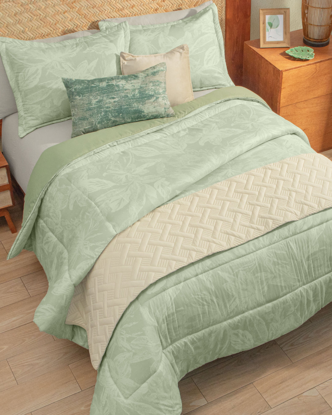 A neatly made bed features the Juego de Edredón Súper Suave Capri by Intima Hogar, showcasing light green floral bedding, a cream cubrecama, and three fundas decorativas accenting the pillows. Beside the bed is a wooden nightstand topped with a picture frame, plant, and a green dish.