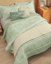 A neatly made bed features the Juego de Edredón Súper Suave Capri by Intima Hogar, showcasing light green floral bedding, a cream cubrecama, and three fundas decorativas accenting the pillows. Beside the bed is a wooden nightstand topped with a picture frame, plant, and a green dish.