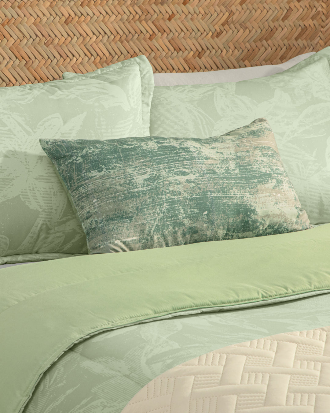 A bed dressed with the Intima Hogar Juego de Edredón Súper Suave Capri, featuring light green floral bedding and a textured green decorative pillow, is accented by a beige quilted cubrecama throw at the foot. The headboard showcases a natural wood tone woven in a herringbone pattern.
