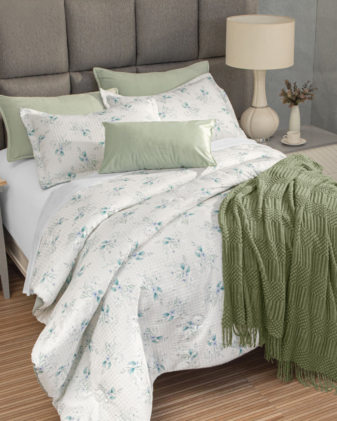 A neatly made bed with the Juego de Edredón Luxury Celeste by Intima Hogar, complemented by green decorative pillow covers and a textured green throw blanket, creates a cozy atmosphere in the dormitorio. A bedside table holds a lamp, a cup, and a small vase with dried flowers.