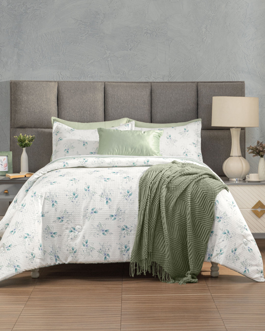 A neatly made bed featuring the Juego de Edredón Luxury Celeste by Intima Hogar, complemented by a green throw blanket, matching green accent pillow, and a gray padded headboard sets the tone for this inviting dormitorio. Nightstands with lamps, a clock, and small decor items — along with stylish fundas decorativas — are placed on either side to complete the cozy look.