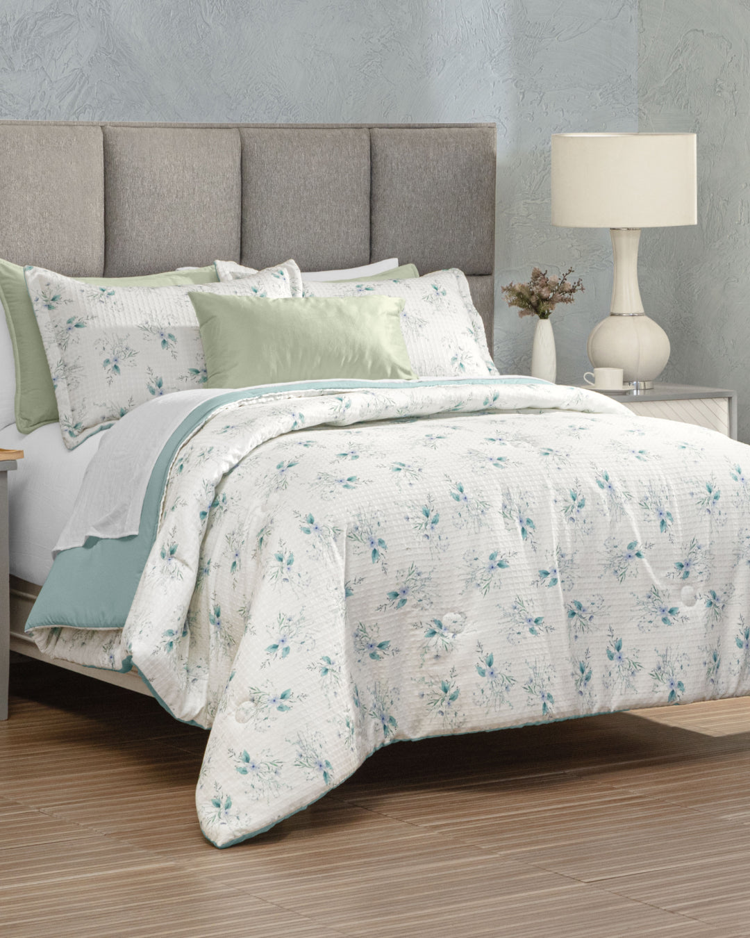 A neatly made bed with a light gray upholstered headboard features the Juego de Edredón Luxury Celeste by Intima Hogar, showcasing white and blue floral bedding, green fundas decorativas accent pillows, and a white lamp on a nightstand—all set against a textured gray wall and wood floor to create an inviting dormitorio.