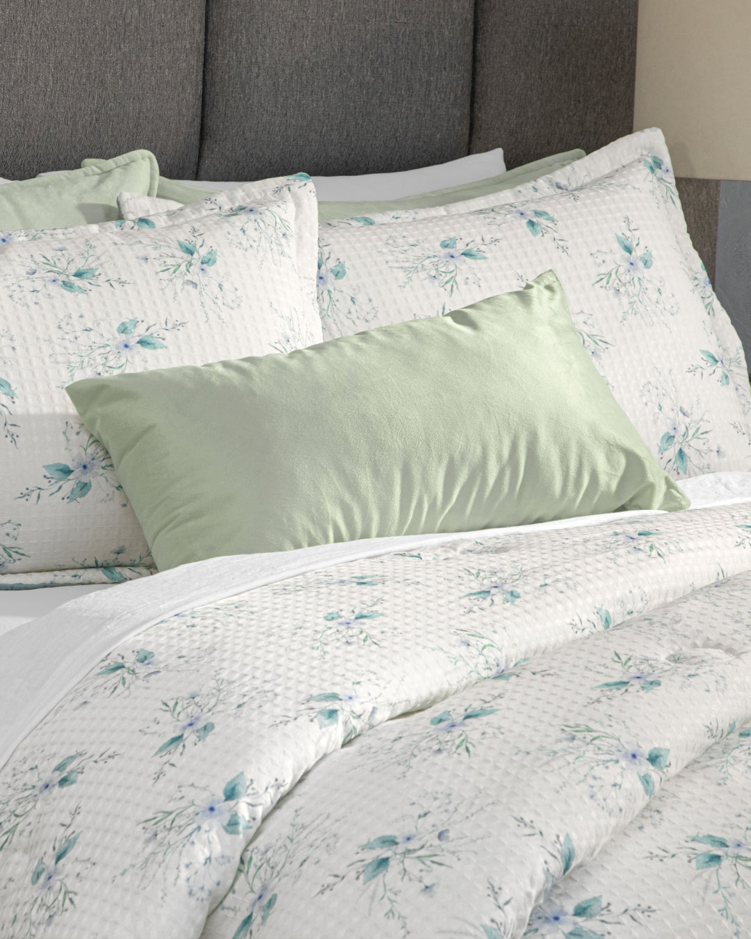 A neatly made dormitorio bed featuring the Juego de Edredón Luxury Celeste by Intima Hogar, with its textured white edredón and pillow shams adorned with a blue floral pattern, complemented by fundas decorativas and a solid light green pillow at the front, all set against a gray upholstered headboard.