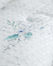 Close-up of a white textured fabric from the Juego de Edredón Luxury Celeste by Intima Hogar, featuring a subtle, light blue and green floral pattern. The soft, slightly raised surface suggests it is part of this decorative bedding set for the dormitorio.