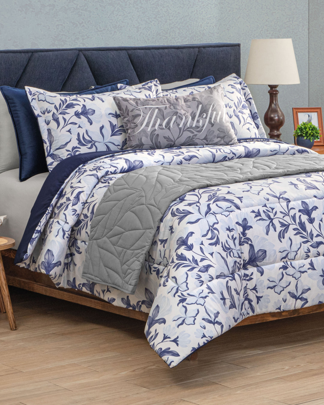 A neatly made bed features the Intima Hogar Juego de Edredón Súper Suave Iris with its blue and white floral design, paired with navy pillows, a Thankful decorative pillow, a dark headboard, and a lamp and framed photo on a wooden nightstand.