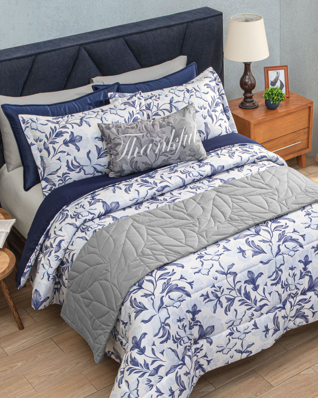 A neatly made bed featuring the Intima Hogar Juego de Edredón Súper Suave Iris with blue and white floral bedding, complemented by a gray edredón and a Thankful decorative pillow with matching fundas decorativas. Beside the bed is a wooden nightstand holding a lamp, framed photo, and clock.
