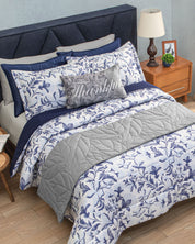 A neatly made bed featuring the Intima Hogar Juego de Edredón Súper Suave Iris with blue and white floral bedding, complemented by a gray edredón and a Thankful decorative pillow with matching fundas decorativas. Beside the bed is a wooden nightstand holding a lamp, framed photo, and clock.