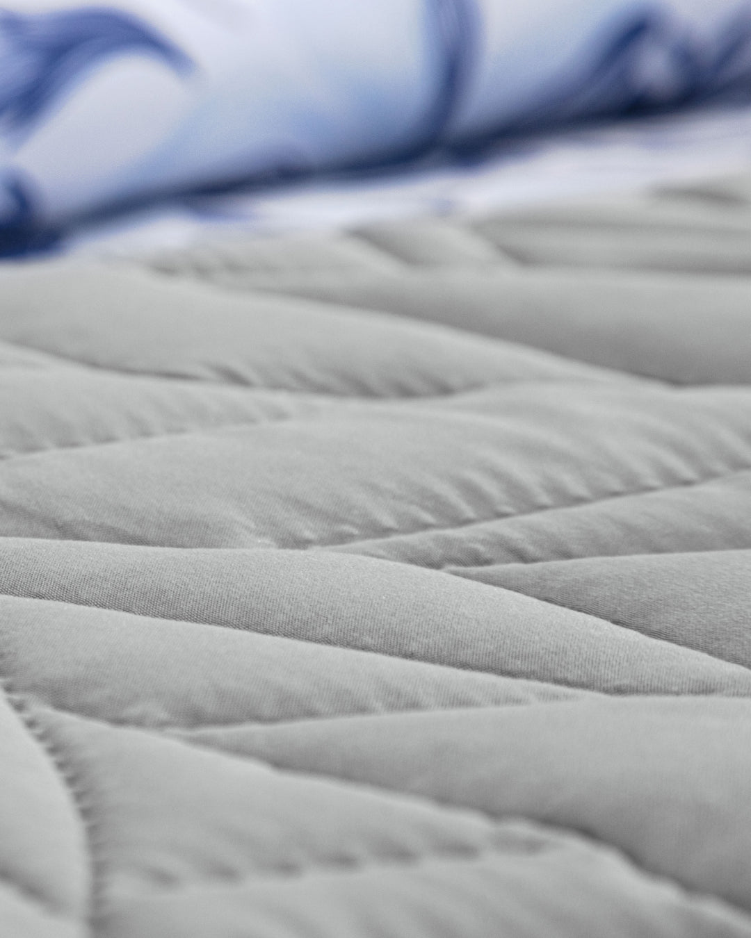 Close-up of a quilted gray mattress surface with visible stitching patterns; in the background, a blurred blue-and-white bedsheet and the elegant Juego de Edredón Súper Suave Iris by Intima Hogar are visible.